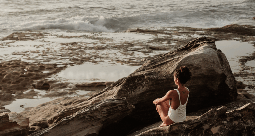 Woman wearing white swimwear, sitting on rocks next to the ocean
